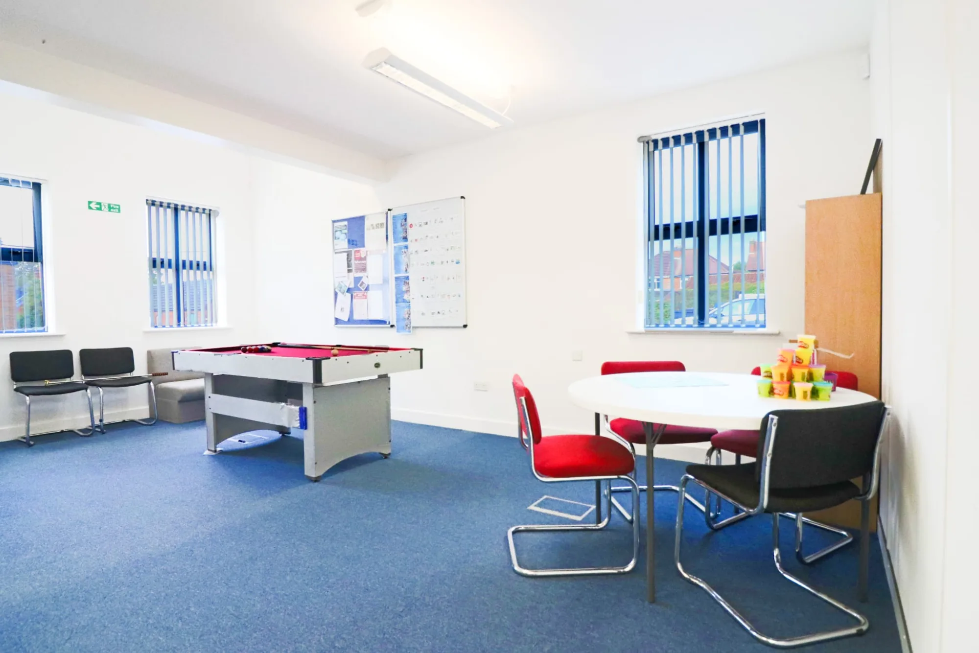 A games room with a red table football table, a whiteboard on the wall and a round white table with red and black chairs and stacking blocks on it.