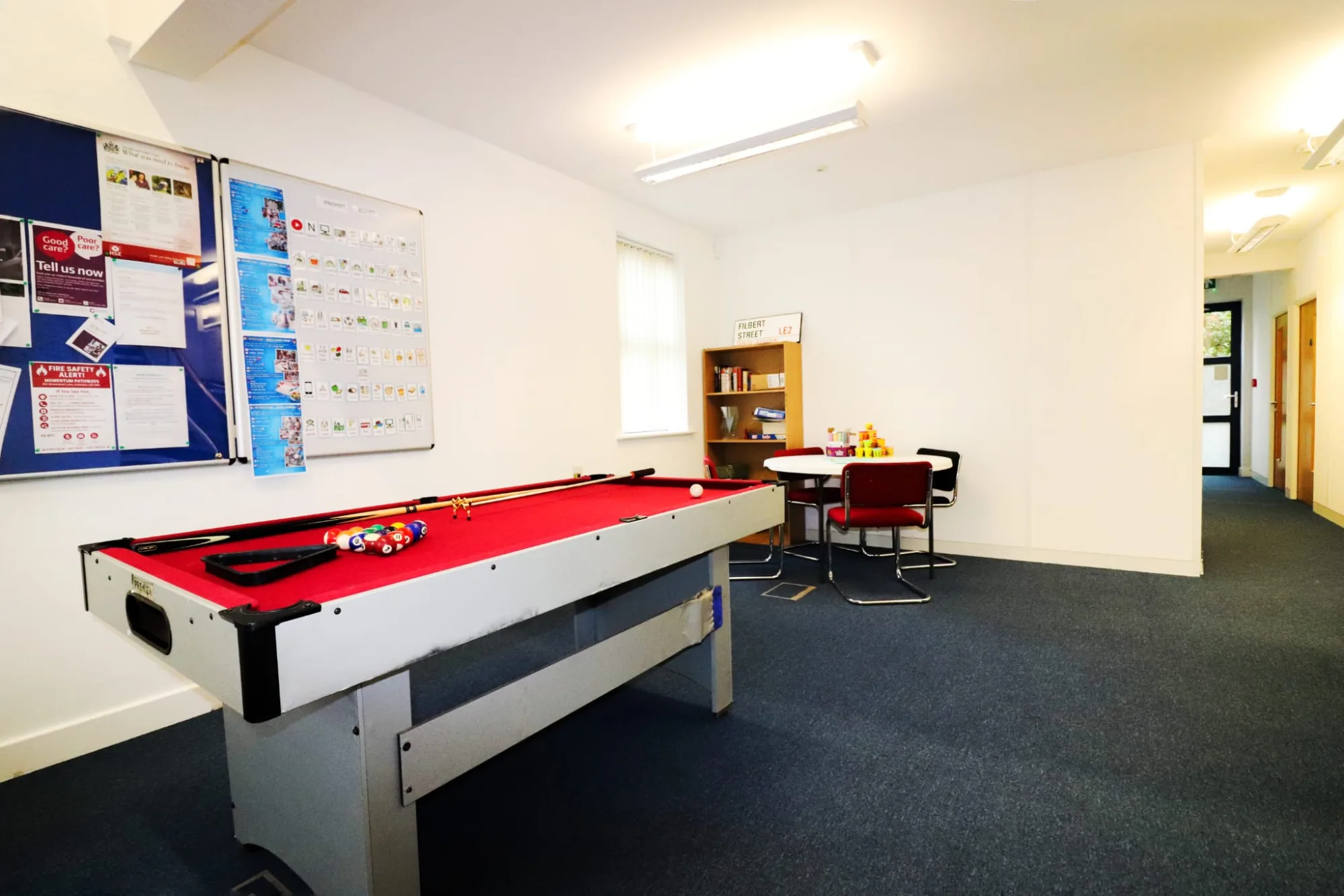 A room with a red baize pool table, notice boards on the wall behind it and a round white table with red chairs and a bookcase beyond.