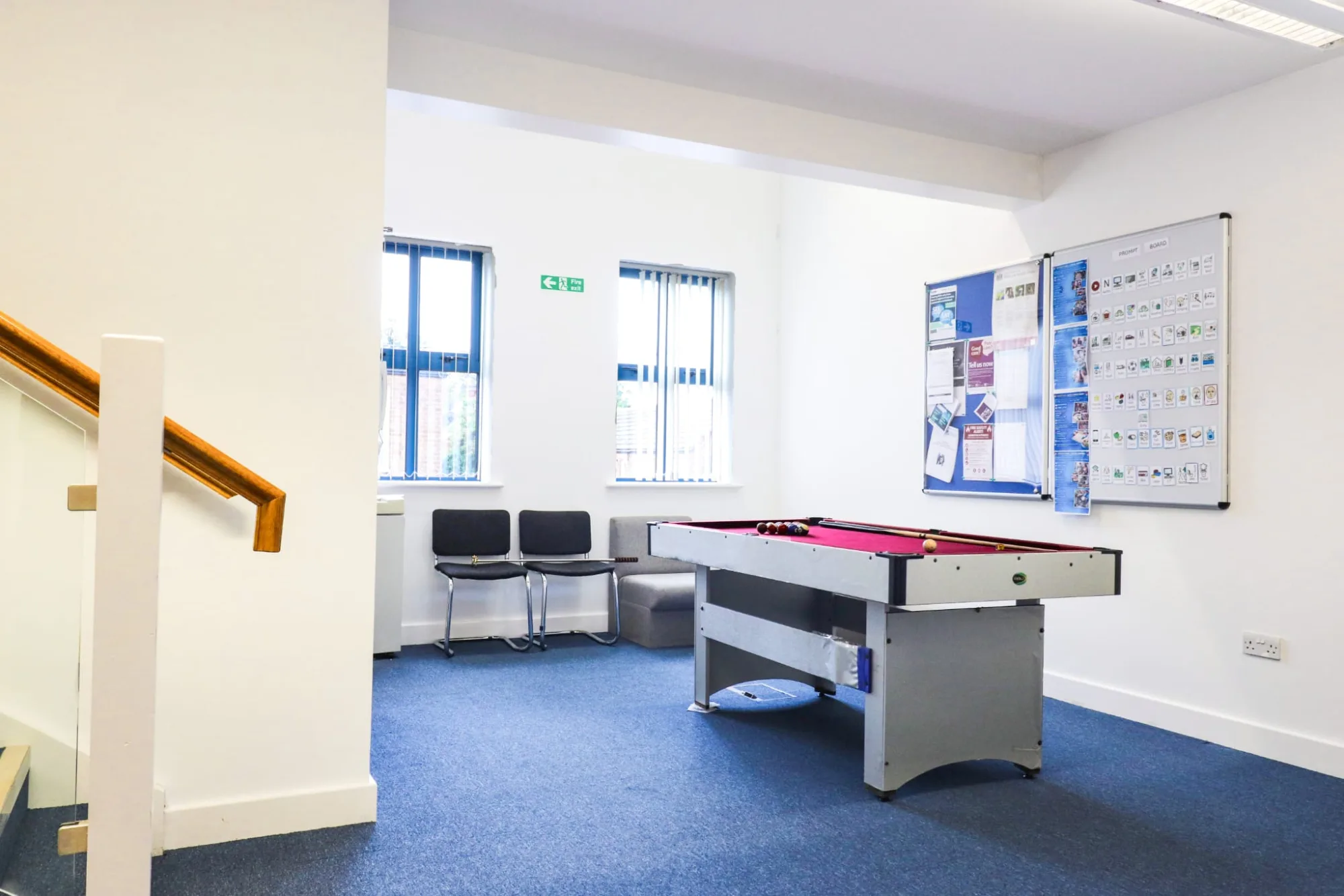 A landing at the top of a staircase with a pool table, chairs against the wall and notice boards beside them.