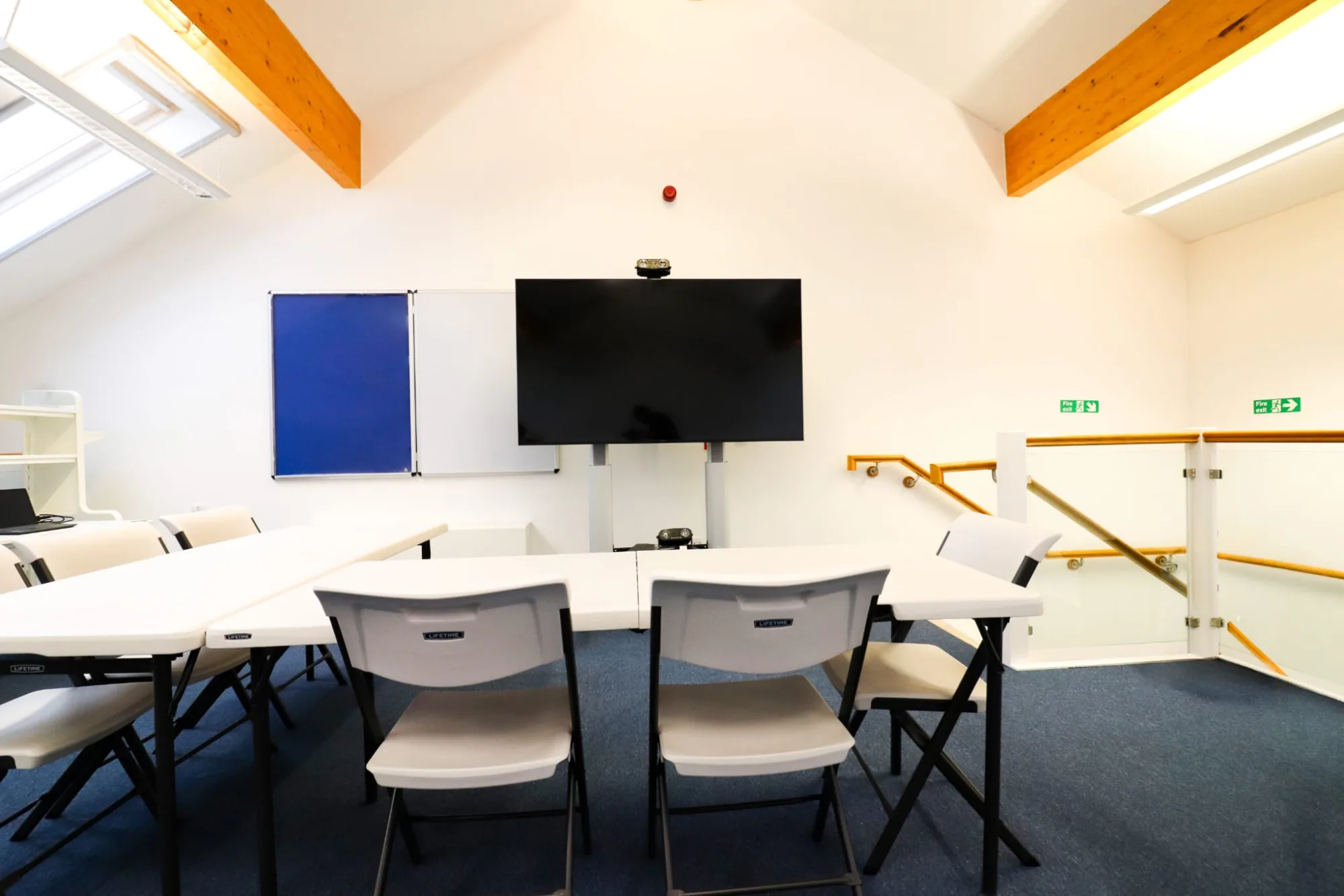 Folding tables and chairs set out facing a large wall-mounted screen, a whiteboard and a pinboard, under a pitched roof with skylights.