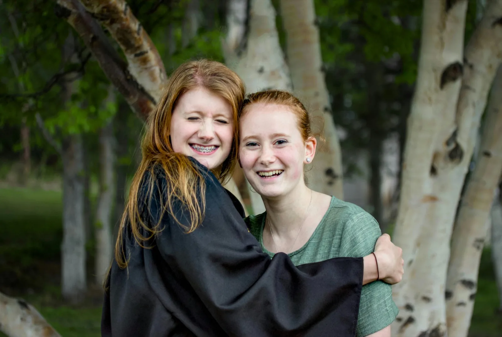 Two young women standing together outdoors among birch trees.